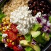 Overhead cottage cheese burrito bowl with brown rice, black beans, corn, romaine lettuce, cherry tomatoes, avocado, red onion, salsa, and a generous scoop of cottage cheese in a black bowl on a rustic wood table.