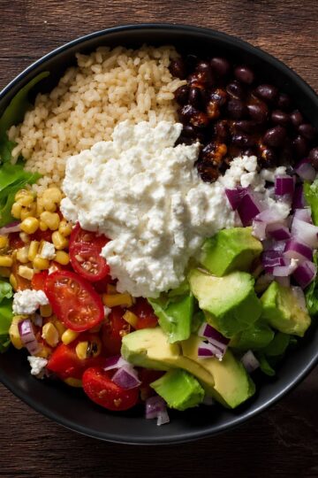 Overhead cottage cheese burrito bowl with brown rice, black beans, corn, romaine lettuce, cherry tomatoes, avocado, red onion, salsa, and a generous scoop of cottage cheese in a black bowl on a rustic wood table.