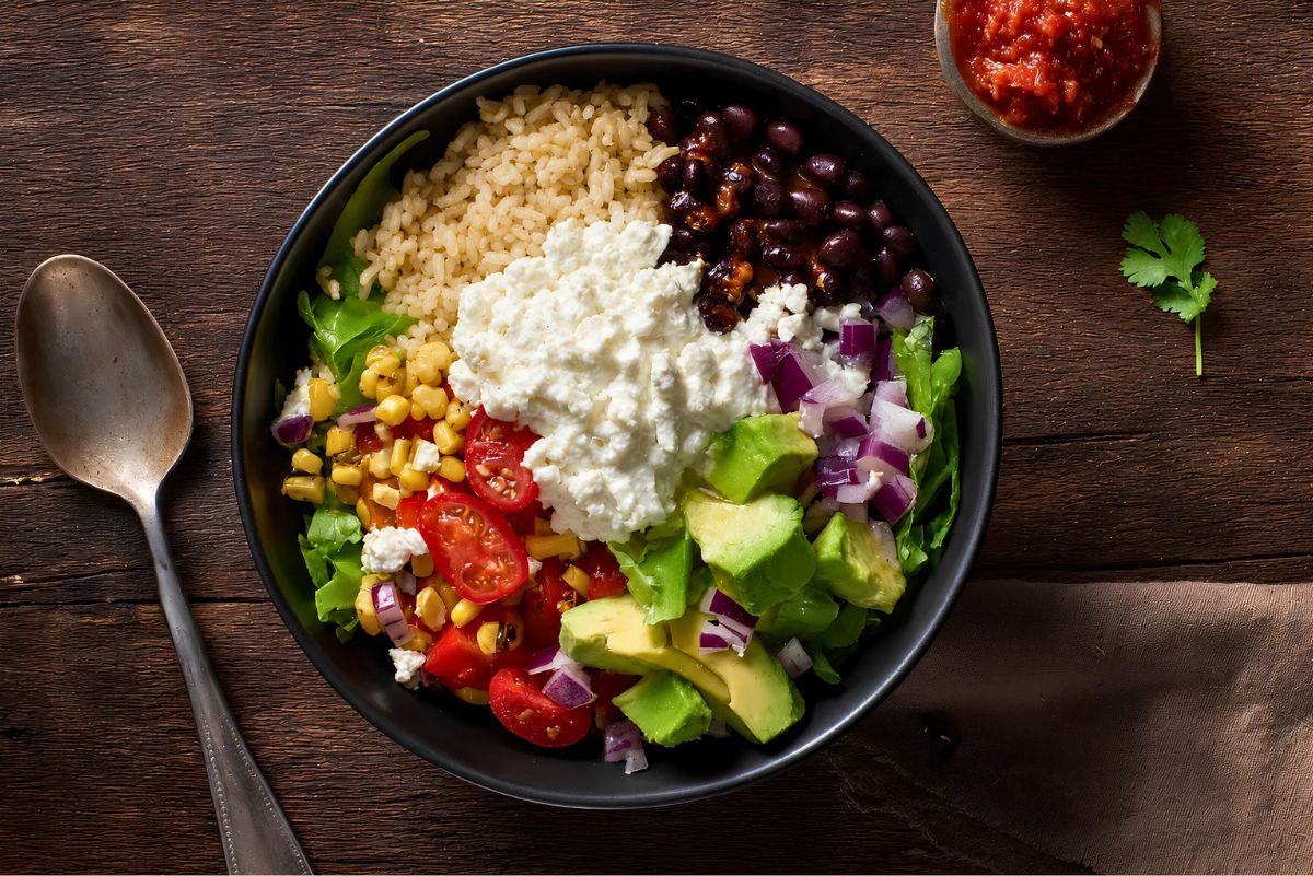 Overhead cottage cheese burrito bowl with brown rice, black beans, corn, romaine lettuce, cherry tomatoes, avocado, red onion, salsa, and creamy cottage cheese in a black bowl on a rustic wood table.