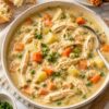 Creamy high protein chicken pot pie soup with shredded chicken, potatoes, carrots, peas, celery, and herbs in a rich creamy broth, served in a large bowl with rustic bread and puff pastry twists nearby.