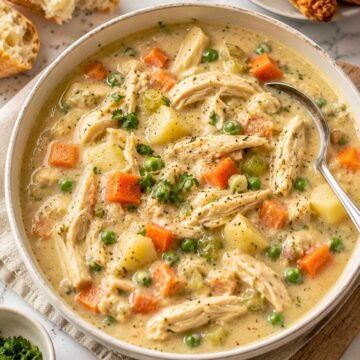 Creamy high protein chicken pot pie soup with shredded chicken, potatoes, carrots, peas, celery, and herbs in a rich creamy broth, served in a large bowl with rustic bread and puff pastry twists nearby.