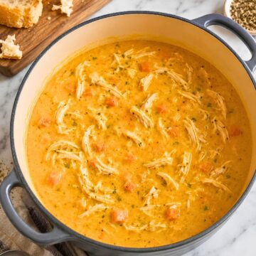 Creamy pumpkin chicken soup with shredded chicken and carrots in a large Dutch oven, served on a marble countertop with sliced crusty bread, herbs, black pepper, and a soup spoon nearby.