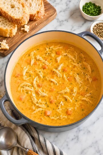 Creamy pumpkin chicken soup with shredded chicken and carrots in a large Dutch oven, served on a marble countertop with sliced crusty bread, herbs, black pepper, and a soup spoon nearby.