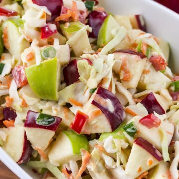 Crunchy apple coleslaw with chunks of green and red apples, shredded cabbage and carrots, diced red bell pepper, and sliced green onion tossed in a creamy dressing and served in a white dish.
