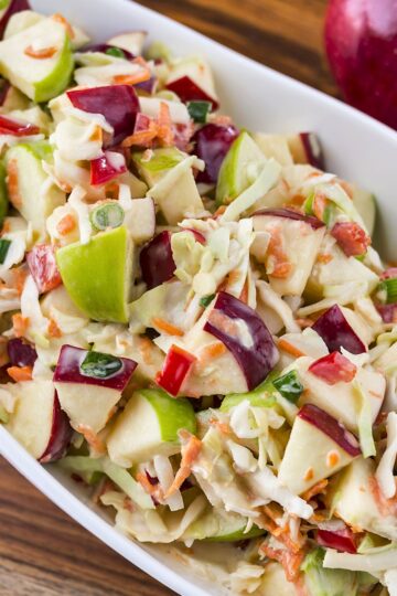 Crunchy apple coleslaw with chunks of green and red apples, shredded cabbage and carrots, diced red bell pepper, and sliced green onion tossed in a creamy dressing and served in a white dish.