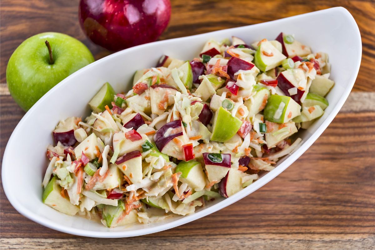 Crunchy apple coleslaw with red and green apples, shredded cabbage and carrots, red bell pepper, and green onion tossed in a creamy dressing. Served in a white oval bowl on a rustic wood table with whole red and Granny Smith apples in the background.