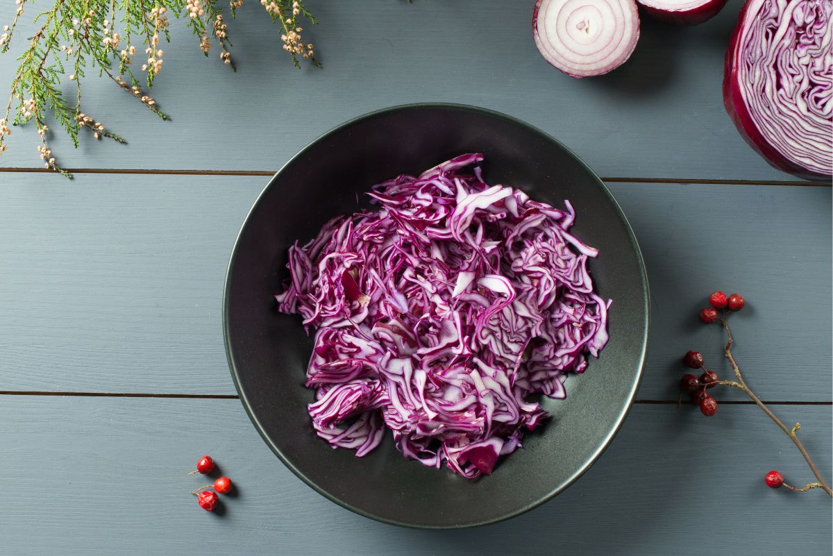 Overhead view of crunchy red cabbage lime slaw in a black bowl, with thinly sliced purple cabbage and red onion. Fresh red cabbage and onion halves surround the bowl on a rustic blue-gray wooden table.