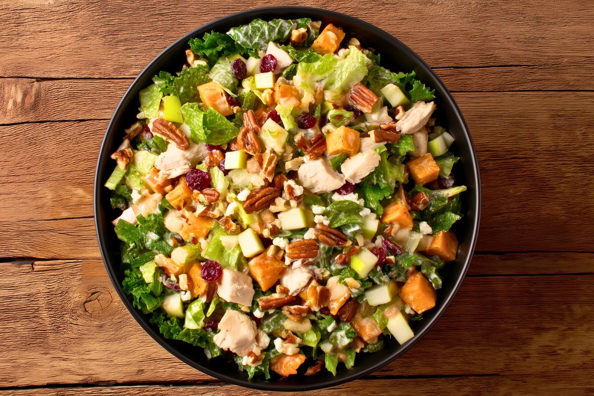 Fall chicken chopped salad with chicken, apples, sweet potatoes, cranberries, pecans, and mixed greens in a black bowl on a wooden table.