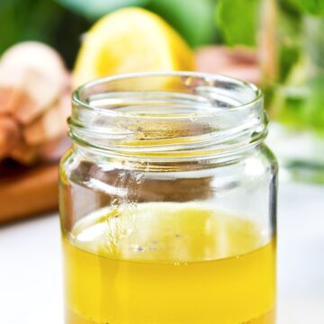 Fresh lemon garlic vinaigrette in a clear glass jar with golden dressing and black pepper, with a lemon, garlic bulb, and fresh herbs softly blurred in the background.