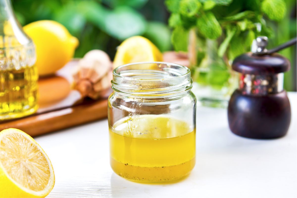 Fresh lemon garlic vinaigrette in a clear glass jar with golden dressing and black pepper, surrounded by fresh lemons, garlic, herbs, olive oil, and a pepper grinder.