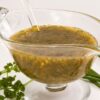 Fresh parsley and chive herb vinaigrette in a clear glass gravy boat with a serving spoon. The green herb dressing has visible seasonings and red pepper flakes, with fresh parsley, chives, and black peppercorns arranged around the dish.
