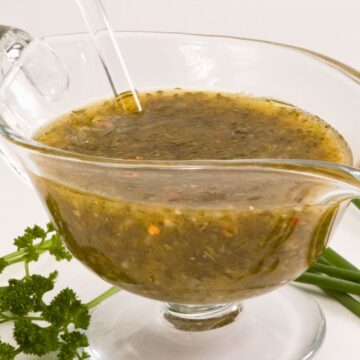 Fresh parsley and chive herb vinaigrette in a clear glass gravy boat with a serving spoon. The green herb dressing has visible seasonings and red pepper flakes, with fresh parsley, chives, and black peppercorns arranged around the dish.
