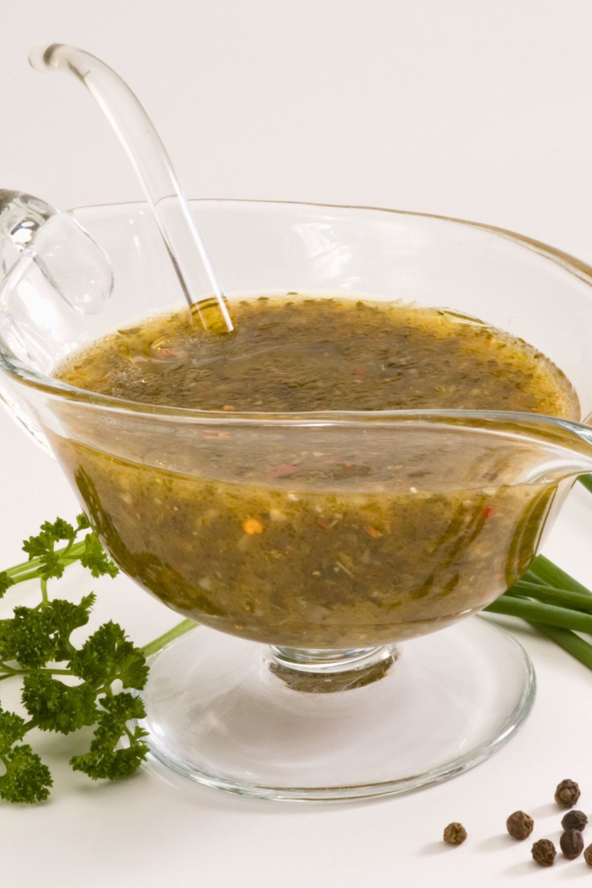 Fresh parsley and chive herb vinaigrette in a clear glass gravy boat with a serving spoon. The green herb dressing has visible seasonings and red pepper flakes, with fresh parsley, chives, and black peppercorns arranged around the dish.