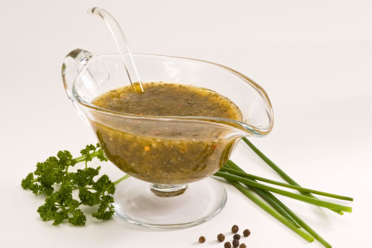 Fresh parsley and chive herb vinaigrette in a clear glass gravy boat with a serving spoon. Fresh parsley and long chive stems surround the homemade salad dressing, with a few black peppercorns scattered on the white surface.