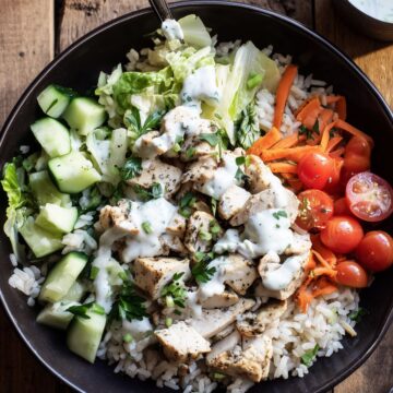 Garlic herb chicken and brown rice bowl with romaine, cucumber, carrots, cherry tomatoes, parsley, and creamy yogurt sauce, served with extra dressing and lemon.