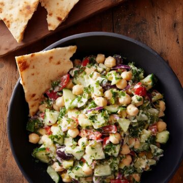 Grinder bean scoopable salad with chickpeas, cucumber, tomatoes, red onion, olives, chopped greens, and creamy dressing in a black bowl. Pita chips sit in the bowl and on a wooden board.