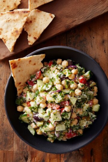 Grinder bean scoopable salad with chickpeas, cucumber, tomatoes, red onion, olives, chopped greens, and creamy dressing in a black bowl. Pita chips sit in the bowl and on a wooden board.