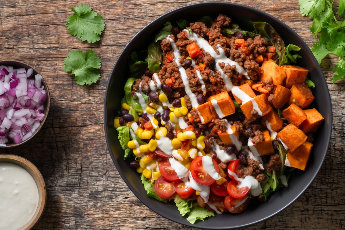 Ground beef and sweet potato bowl with seasoned ground beef, roasted sweet potatoes, black beans, corn, cherry tomatoes, leafy greens, and creamy dressing, served in a black bowl with red onion, cilantro, and extra dressing on the side.