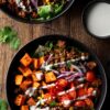 Ground beef and sweet potato bowls with roasted sweet potatoes, seasoned ground beef, black beans, corn, cherry tomatoes, red onion, leafy greens, cilantro, and creamy dressing, served in black bowls on a rustic wooden table.
