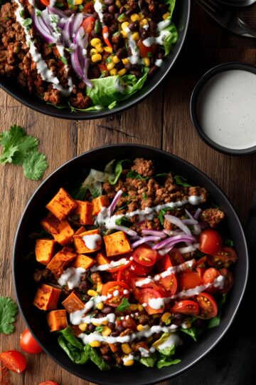 Ground beef and sweet potato bowls with roasted sweet potatoes, seasoned ground beef, black beans, corn, cherry tomatoes, red onion, leafy greens, cilantro, and creamy dressing, served in black bowls on a rustic wooden table.