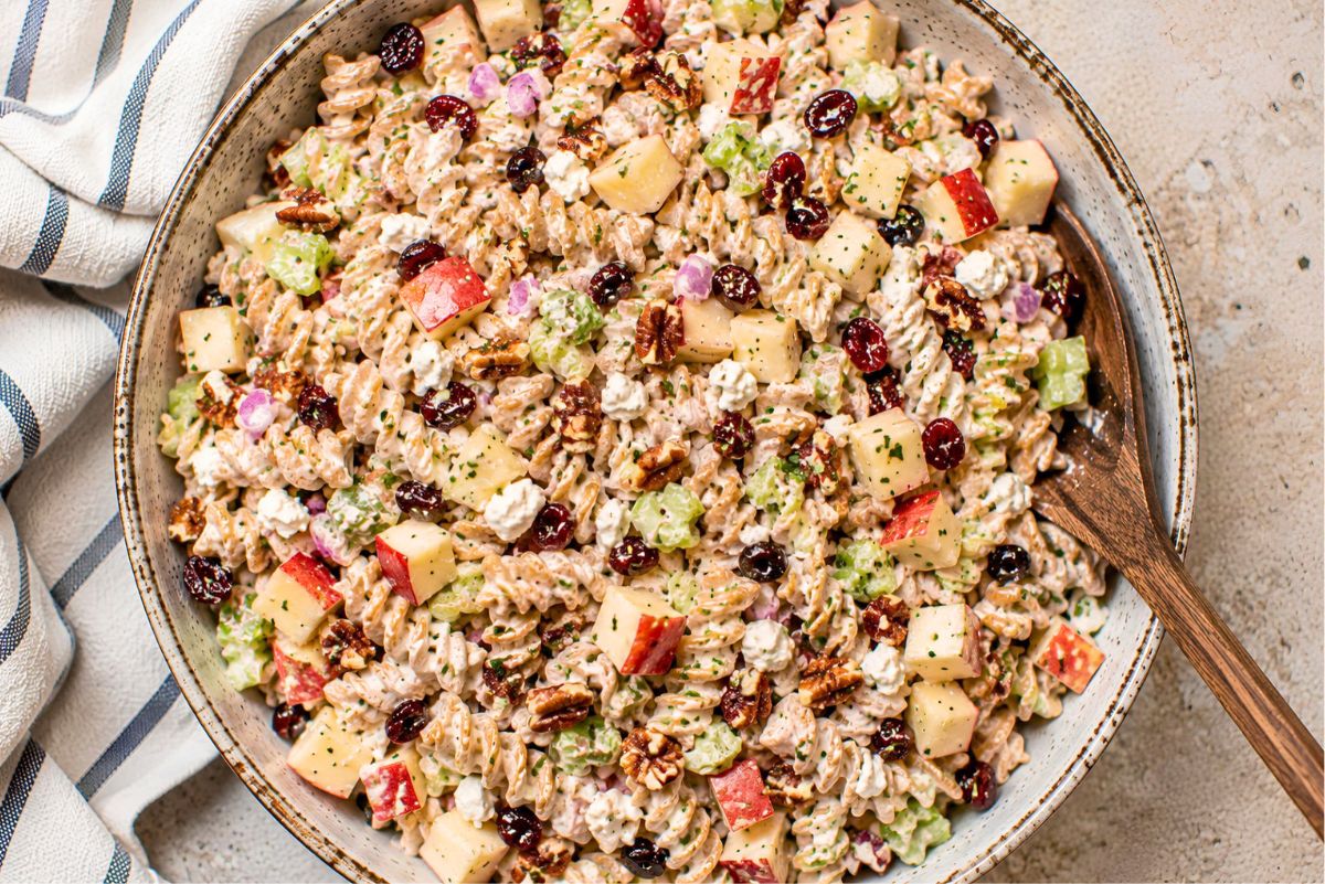 Healthy apple pecan pasta salad with whole wheat rotini, chopped red apples, celery, dried cranberries, pecans, red onion, feta cheese, and herbs in a creamy dressing, served in a large rustic bowl with a wooden spoon.