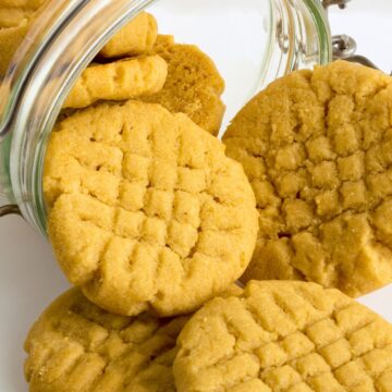 Golden peanut butter chickpea cookies with crisscross fork marks spilling from an open glass storage jar onto a white surface.