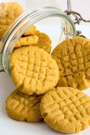 Golden peanut butter chickpea cookies with crisscross fork marks spilling from an open glass storage jar onto a white surface.