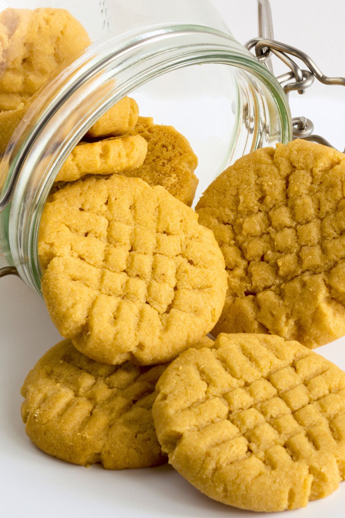 Golden peanut butter chickpea cookies with crisscross fork marks spilling from an open glass storage jar onto a white surface.