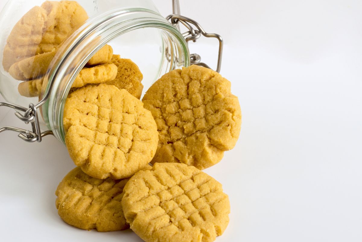 Peanut butter chickpea cookies with crisscross fork marks spilling from an open glass storage jar onto a white surface, with copy space on the right.