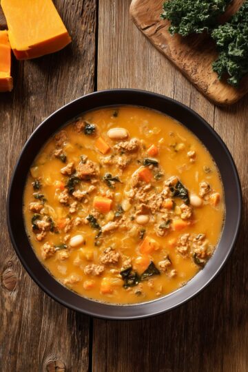 Overhead view of butternut squash turkey soup in a black bowl with ground turkey, kale, carrots, white beans, and a rich orange broth on rustic wood.
