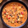 Overhead view of creamy chicken burrito soup with chicken, black beans, corn, tomatoes, and bell peppers in a black bowl on rustic wood, with lime and chopped red pepper nearby.
