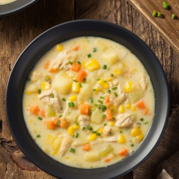 Creamy high protein chicken corn chowder with chicken, corn, potatoes, carrots, celery, and chives in a black bowl on a rustic wooden table. A second bowl, corn cob, and spoon sit nearby.