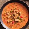 Overhead view of high protein chicken fajita soup with chicken, bell peppers, corn, black beans, and tomatoes in a creamy red broth, served in a black bowl on rustic wood.