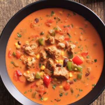 Overhead view of high protein chicken fajita soup with chicken, bell peppers, corn, black beans, and tomatoes in a creamy red broth, served in a black bowl on rustic wood.