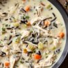 High protein creamy chicken and wild rice soup with shredded chicken, wild rice, carrots, celery, herbs, and vegetables in a rich creamy broth, served in a dark bowl on a rustic wooden table.