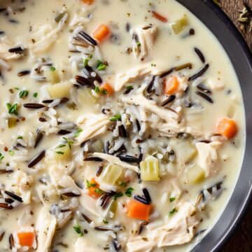 High protein creamy chicken and wild rice soup with shredded chicken, wild rice, carrots, celery, herbs, and vegetables in a rich creamy broth, served in a dark bowl on a rustic wooden table.