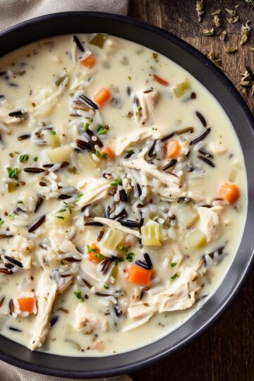 High protein creamy chicken and wild rice soup with shredded chicken, wild rice, carrots, celery, herbs, and vegetables in a rich creamy broth, served in a dark bowl on a rustic wooden table.