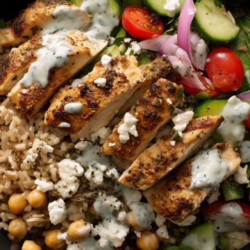 High protein Greek chicken bowl with seasoned grilled chicken, brown rice, chickpeas, cucumber, cherry tomatoes, red onion, feta cheese, leafy greens, and creamy Greek yogurt sauce in a black bowl.