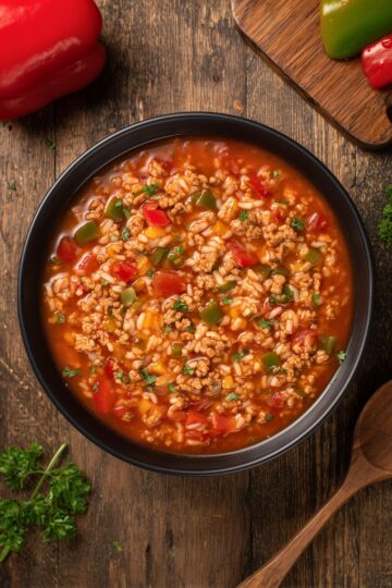 Overhead view of stuffed pepper soup with ground turkey, rice, tomatoes, and colorful bell peppers in a black bowl, surrounded by whole peppers, parsley, and a wooden spoon.