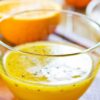 Close-up of homemade orange black sesame vinaigrette in a clear glass bowl, with fresh orange halves in the background.