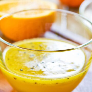 Close-up of homemade orange black sesame vinaigrette in a clear glass bowl, with fresh orange halves in the background.