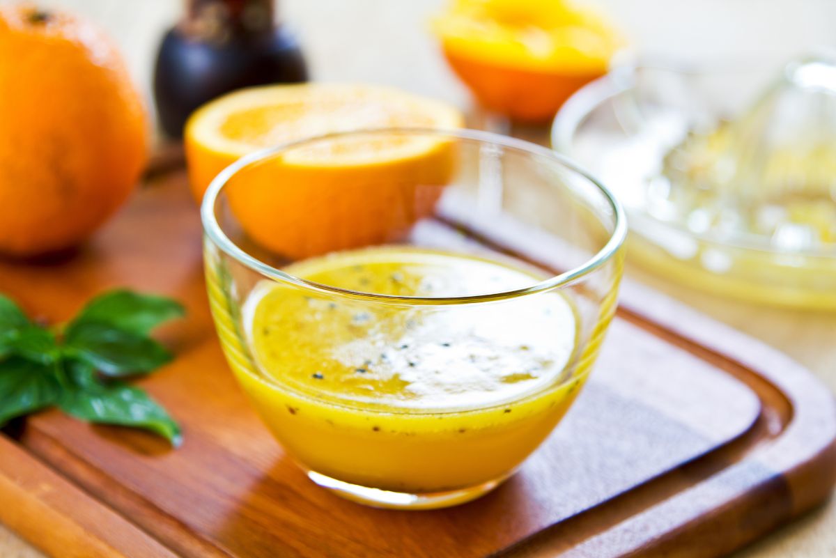 Homemade orange black sesame vinaigrette in a clear glass bowl on a wooden board, surrounded by fresh oranges, basil, and a citrus juicer.