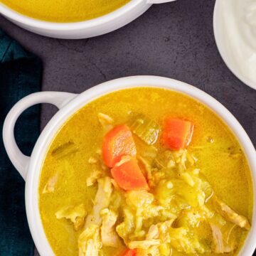 Homestyle chicken mulligatawny soup in white bowls with a golden curry broth, shredded chicken, carrots, rice, and vegetables, served beside a small bowl of creamy white topping on a dark stone surface.