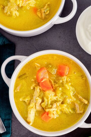 Homestyle chicken mulligatawny soup in white bowls with a golden curry broth, shredded chicken, carrots, rice, and vegetables, served beside a small bowl of creamy white topping on a dark stone surface.