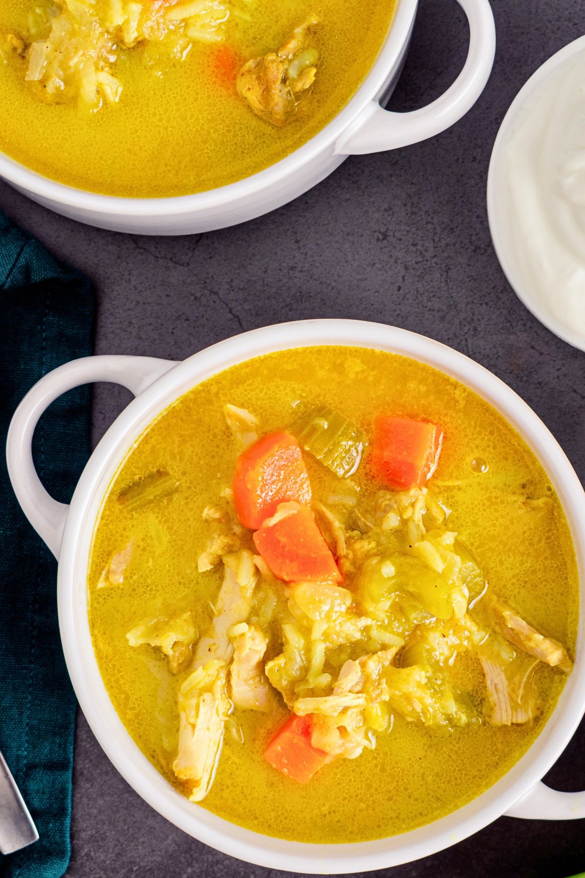 Homestyle chicken mulligatawny soup in white bowls with a golden curry broth, shredded chicken, carrots, rice, and vegetables, served beside a small bowl of creamy white topping on a dark stone surface.