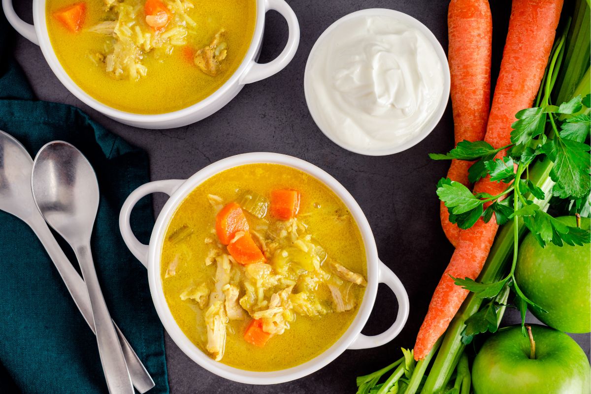 Homestyle chicken mulligatawny soup with golden curry broth, shredded chicken, carrots, rice, and vegetables in white bowls, surrounded by fresh carrots, celery, green apples, parsley, and a bowl of creamy white topping.