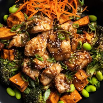 Overhead honey garlic chicken sweet potato bowl with glazed chicken, roasted sweet potatoes, broccoli, shredded carrots, edamame, green onions, and sesame seeds in a black bowl on a rustic wood table.
