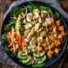 Overhead view of a honey mustard chicken sweet potato bowl with roasted vegetables, fresh greens, cucumber, carrots, pumpkin seeds, and creamy sauce.