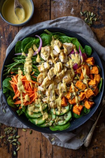 Overhead view of a honey mustard chicken sweet potato bowl with roasted vegetables, fresh greens, cucumber, carrots, pumpkin seeds, and creamy sauce.