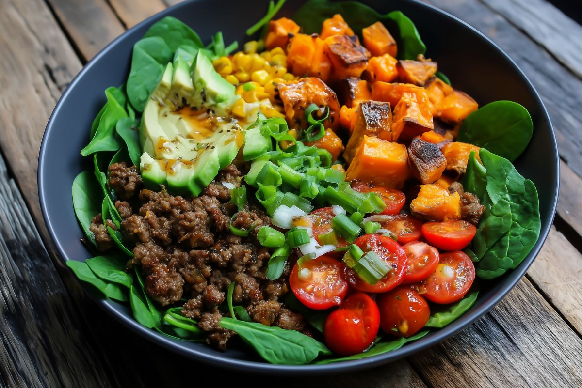 Hot honey beef sweet potato bowl with seasoned ground beef, roasted sweet potatoes, avocado, corn, spinach, cherry tomatoes, and green onions in a black bowl on a rustic wood table.